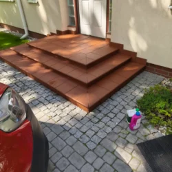 Szerokie schody wejściowe z rudobrązowymi płytkami przed białymi drzwiami.