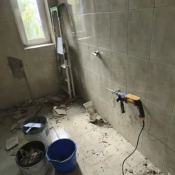 Skuwanie płytek w łazience z użyciem młota udarowego, z wiadrami i gruzem na podłodze.