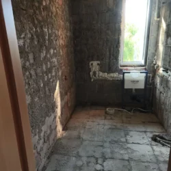 Łazienka po usunięciu okładzin, z odsłoniętymi ścianami i stelażem WC pod oknem.