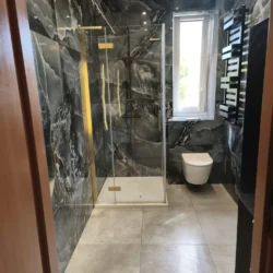 Widok od wejścia na kabinę prysznicową, WC pod oknem i czarny grzejnik.