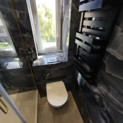 Wisząca miska WC pod oknem i czarny grzejnik obok kabiny prysznicowej.